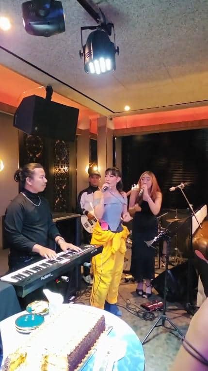 Beberapa orang bernyanyi dan bermain musik di sebuah ruangan dengan pencahayaan hangat saat perayaan ulang tahun, dengan kue di meja depan.