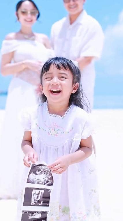 Nadlyne tersenyum lebar sambil memegang hasil USG, dengan dua orang dewasa berdiri di belakangnya di pantai berpasir putih.