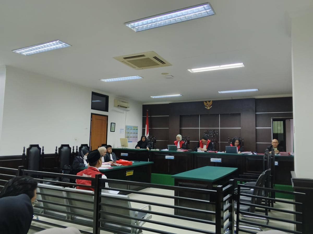 Suasana sidang di ruang pengadilan dengan majelis hakim mengenakan toga merah dan terdakwa duduk di kursi depan bersama penasihat hukum.