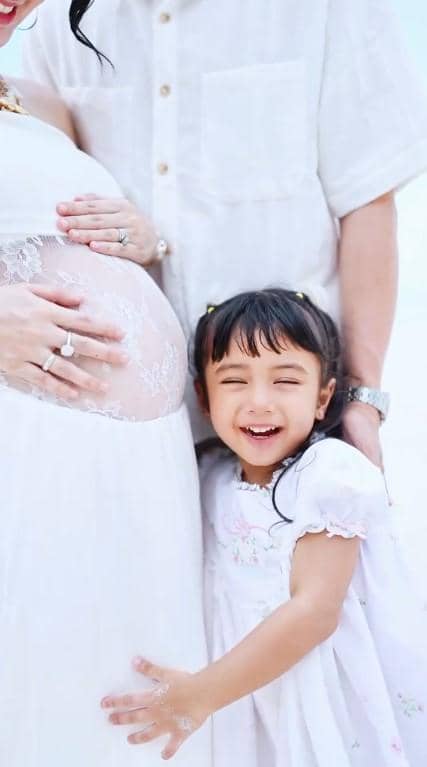 Nadlyne tersenyum sambil memeluk perut ibunya yang sedang hamil, dengan latar suasana cerah dan pakaian berwarna putih.