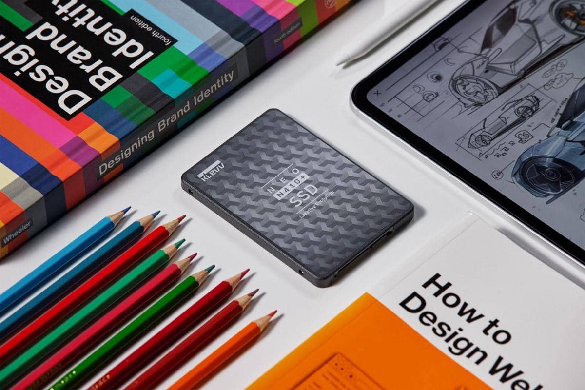 SSD Klevv Neo N410+ berwarna abu-abu ditempatkan di meja bersama buku desain, pensil warna, dan tablet dengan sketsa mobil.