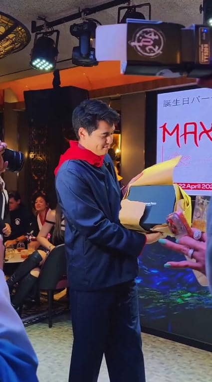 Maxime Bouttier mengenakan jaket biru dan syal merah membuka hadiah di acara ulang tahun di sebuah tempat dengan pencahayaan hangat.