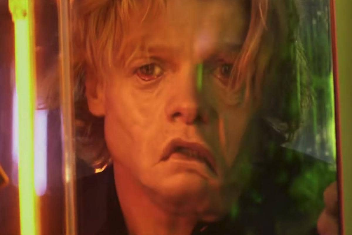 Wajah Matt Hagen dengan ekspresi tegang terlihat melalui pantulan kaca berwarna oranye dan hijau dalam cuplikan teaser film Clayface.