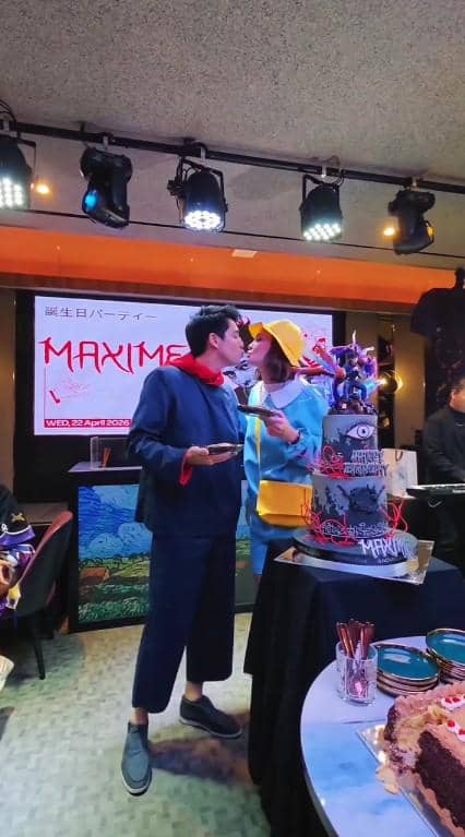 Maxime dan Luna berdiri berdekatan di pesta ulang tahun dengan kue bertingkat bertema unik di atas meja