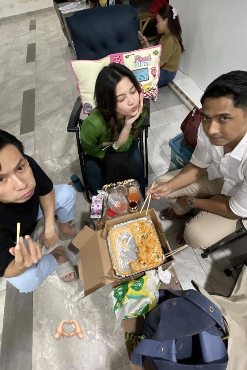 Tiga orang duduk santai di lokasi syuting sambil menikmati makanan dari kotak besar dengan sumpit di atas lantai berubin.