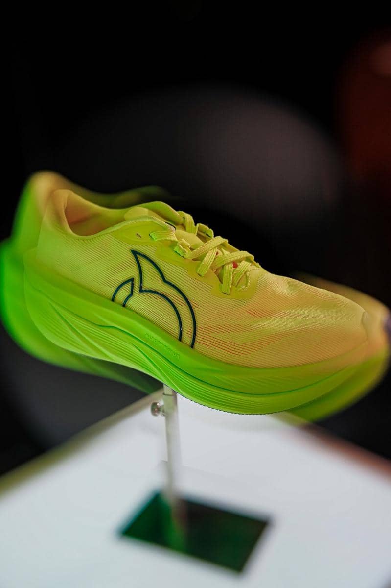 Sepatu olahraga Ortuseight berwarna hijau neon dengan desain modern dan sol tebal dipajang di atas dudukan logam.
