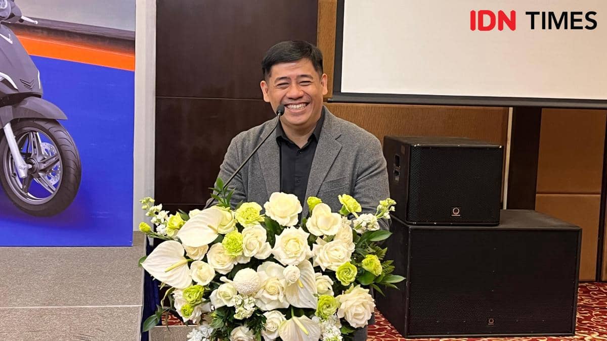 Seorang pria tersenyum saat berbicara di podium dengan hiasan bunga putih dan kuning di depan latar dalam ruangan konferensi.