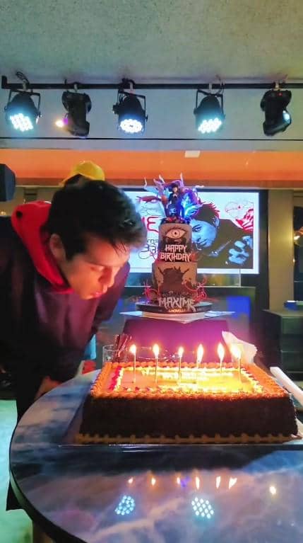 Maxime Bouttier meniup lilin di atas kue ulang tahun cokelat dengan dekorasi pesta dan pencahayaan warna-warni di sebuah ruangan.