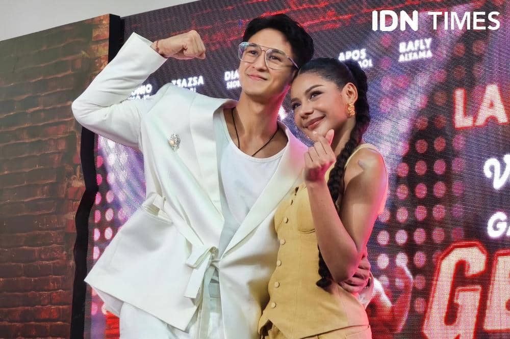 Dua orang berpose bersama di depan latar promosi acara di Jakarta, satu mengenakan setelan putih dan satu mengenakan busana kuning.