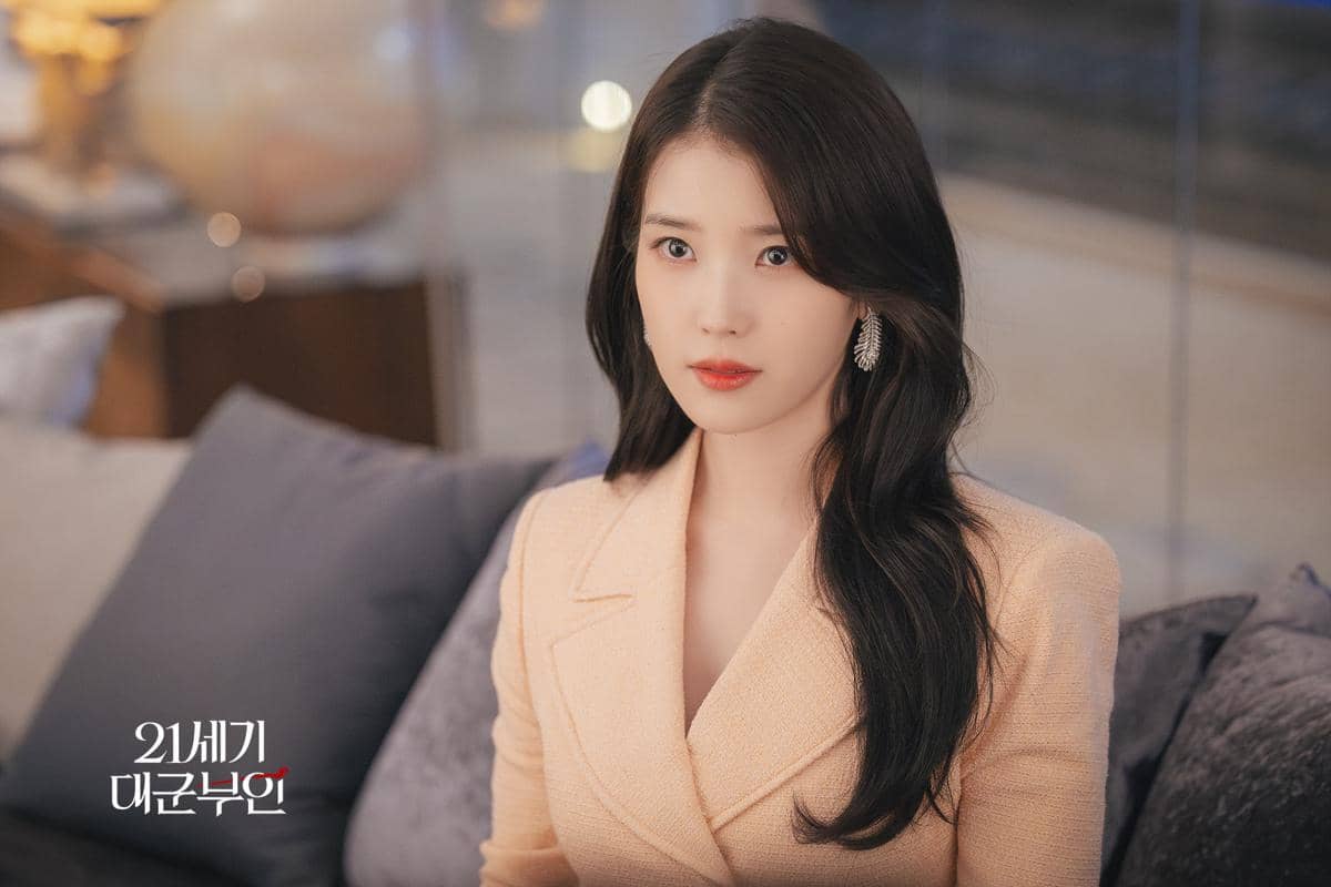 Seorang wanita berambut panjang mengenakan blazer krem duduk di sofa dengan ekspresi serius dalam cuplikan drama Perfect Crown.