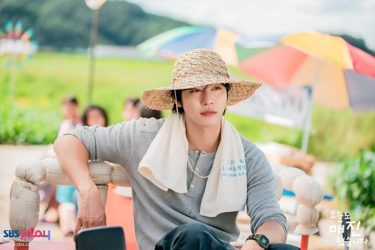Seorang pria mengenakan topi jerami dan handuk di leher duduk santai di luar ruangan dengan latar payung warna-warni dalam adegan drama Sold Out On You.