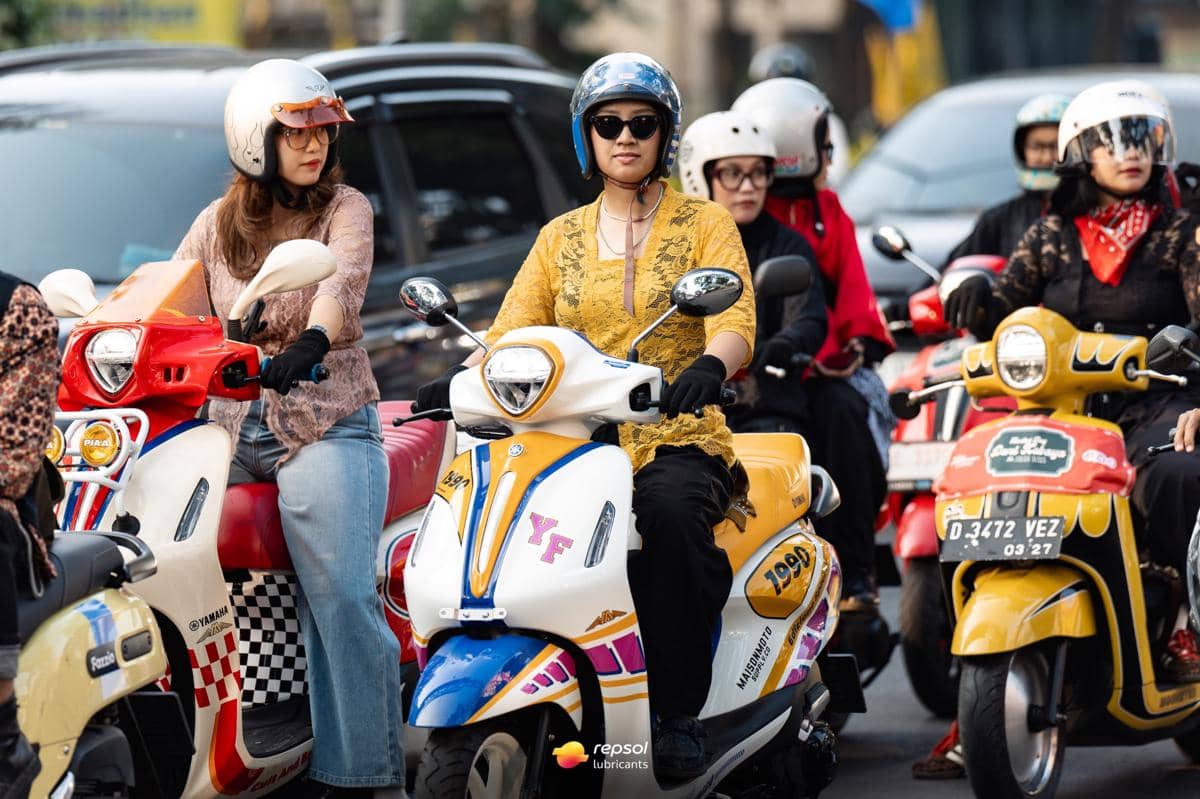 Sekelompok wanita mengenakan kebaya dan helm saat mengendarai skuter berwarna cerah di jalan perkotaan yang ramai.