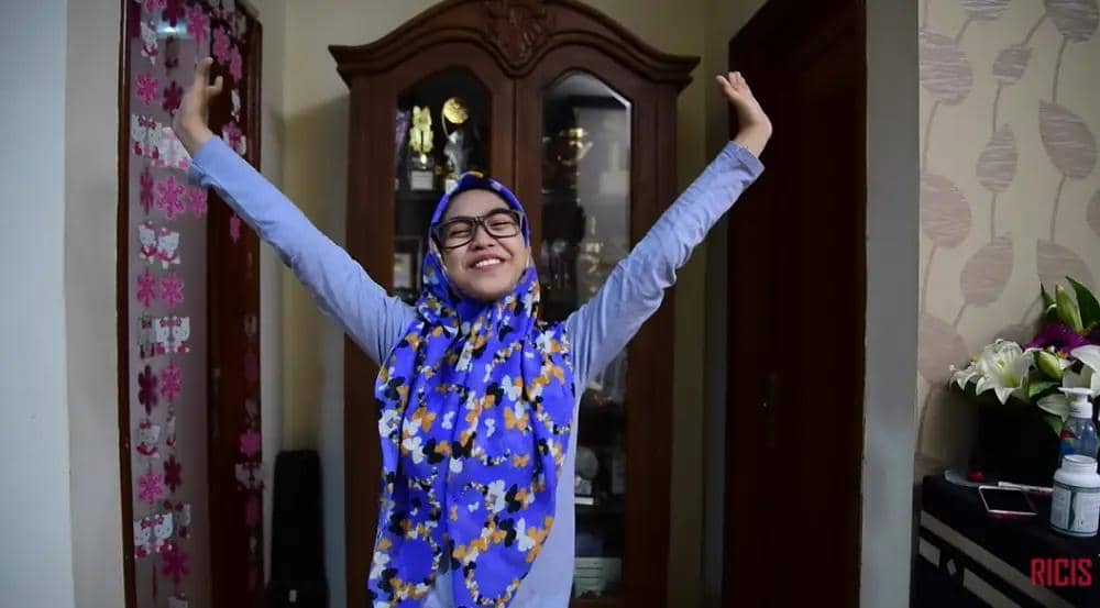 Perempuan berhijab motif bunga biru tersenyum lebar sambil mengangkat kedua tangan di dalam ruangan rumah dengan lemari kayu di belakangnya.