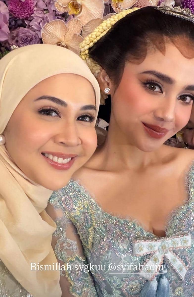 Dua perempuan tersenyum mengenakan busana berwarna pastel berhias payet, dengan latar bunga anggrek ungu dan krem.