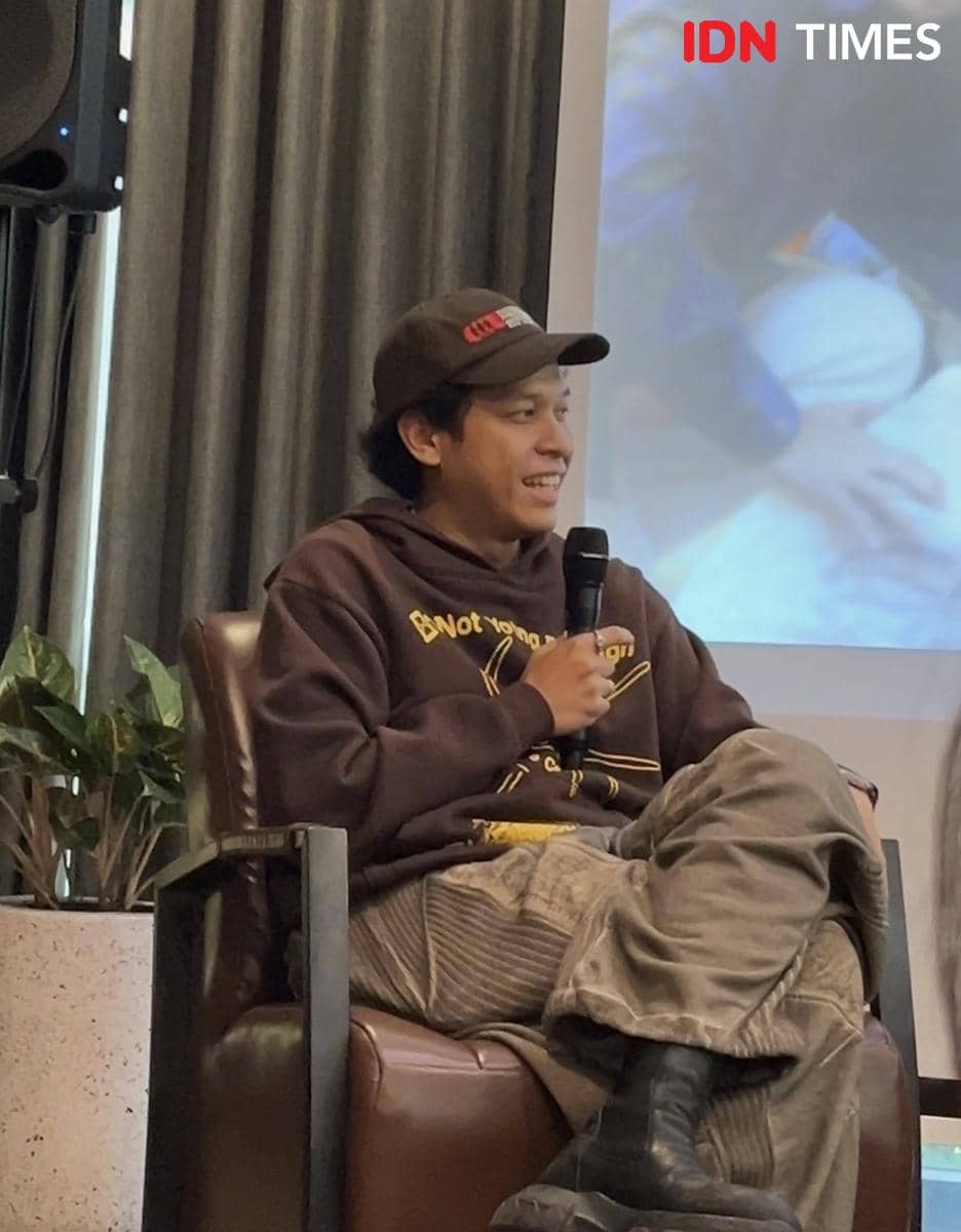 Seorang pria mengenakan hoodie cokelat dan topi hitam sedang berbicara sambil memegang mikrofon dalam wawancara di Jakarta Selatan.