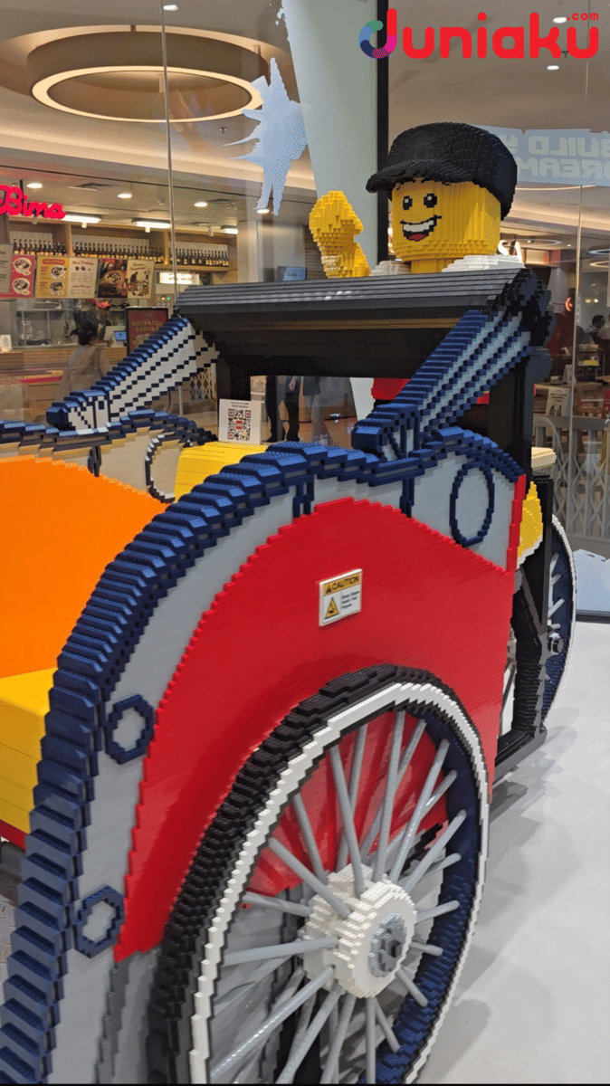 Instalasi becak berwarna cerah yang terbuat dari balok LEGO dengan figur pengemudi tersenyum di LEGO Certified Store Grand Indonesia.