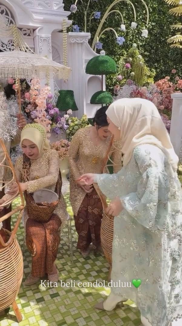 Beberapa wanita mengenakan kebaya dan hijab sedang menikmati suasana pengajian dan siraman dengan latar dekorasi bunga berwarna-warni.