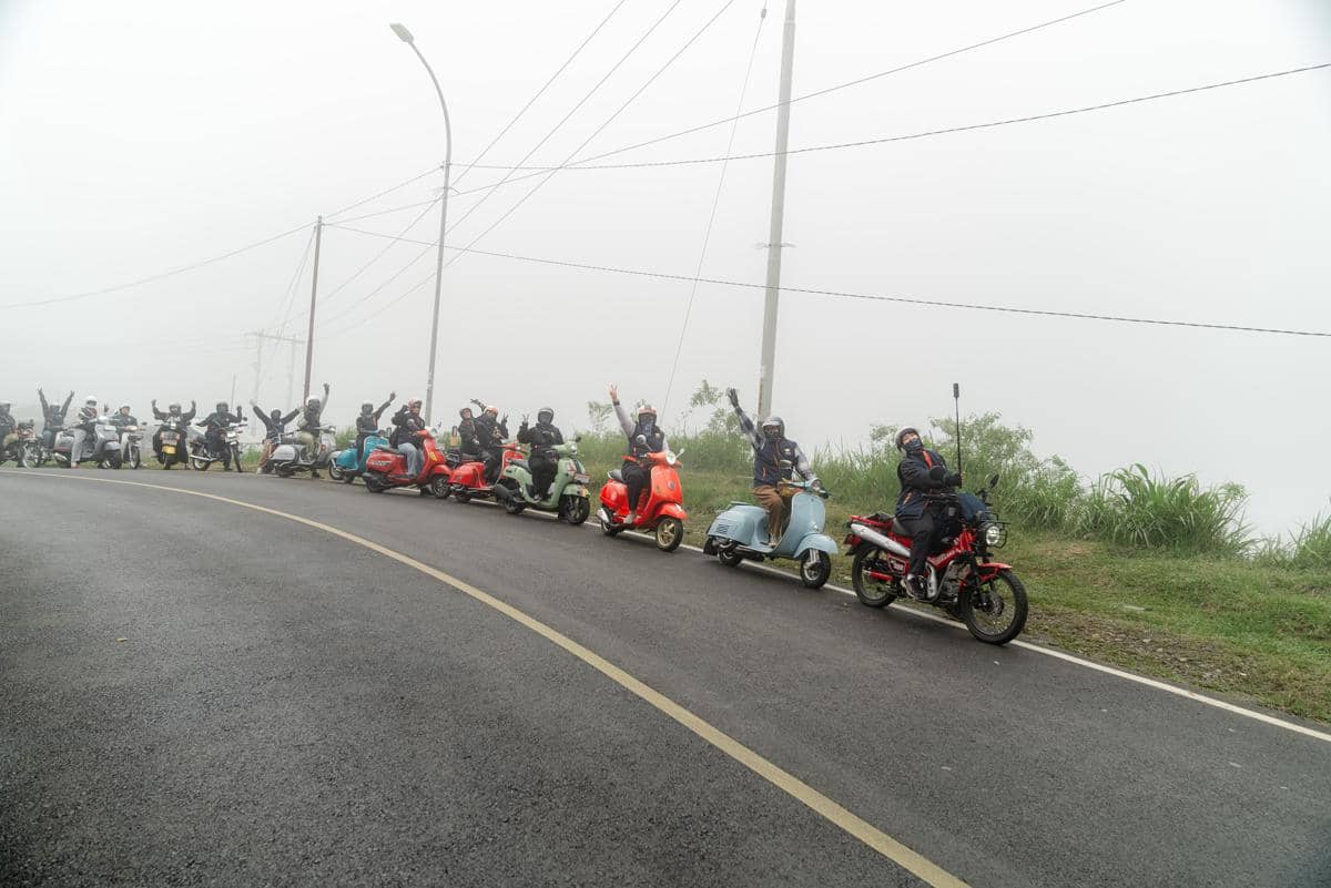 Sekelompok pengendara motor klasik berbaris di jalan beraspal berkabut sambil mengangkat tangan dan mengenakan perlengkapan berkendara lengkap.