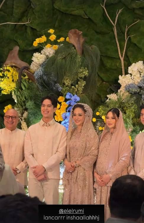 El Rumi berdiri bersama keluarga mengenakan busana tradisional warna krem dalam acara pengajian dan siraman dengan latar bunga berwarna cerah.