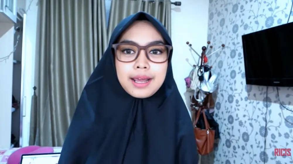Seorang perempuan berhijab hitam dan berkacamata berbicara di dalam kamar dengan latar tirai dan televisi di dinding.
