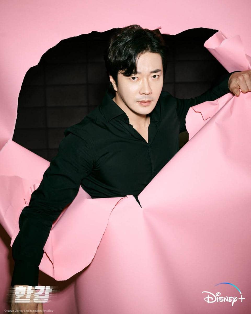 Seorang pria mengenakan kemeja hitam muncul dari sobekan besar kertas berwarna pink dengan latar belakang gelap.