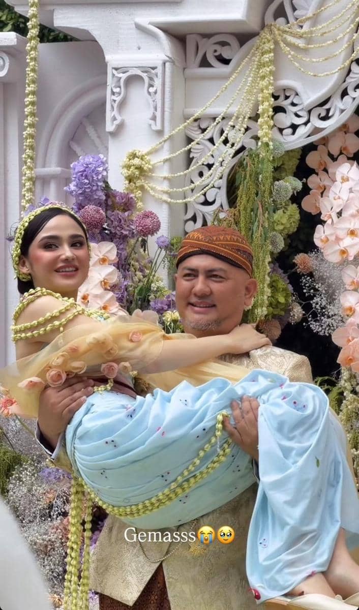 Seorang pria mengenakan pakaian tradisional menggendong seorang wanita berhias busana adat berwarna pastel di acara siraman dengan dekorasi bunga.