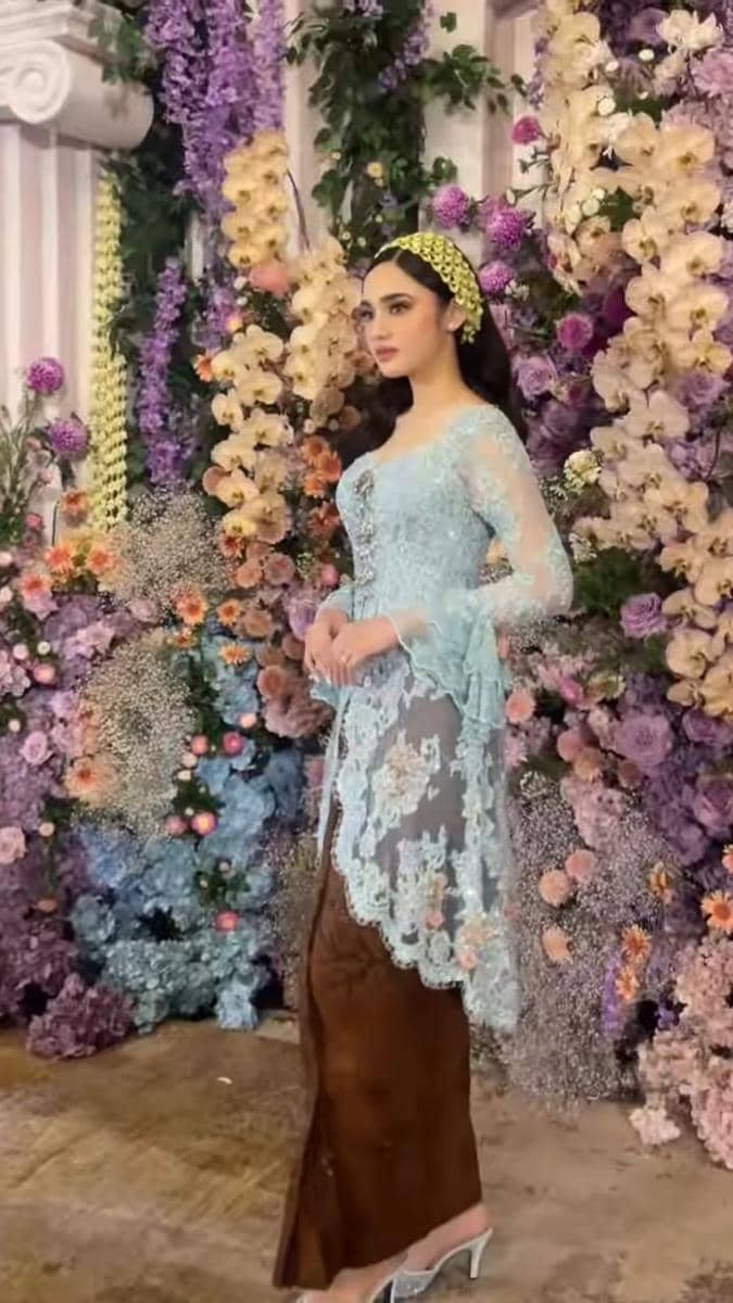 Seorang perempuan mengenakan kebaya biru muda dan kain cokelat berpose di depan dekorasi bunga warna ungu dan krem yang indah.