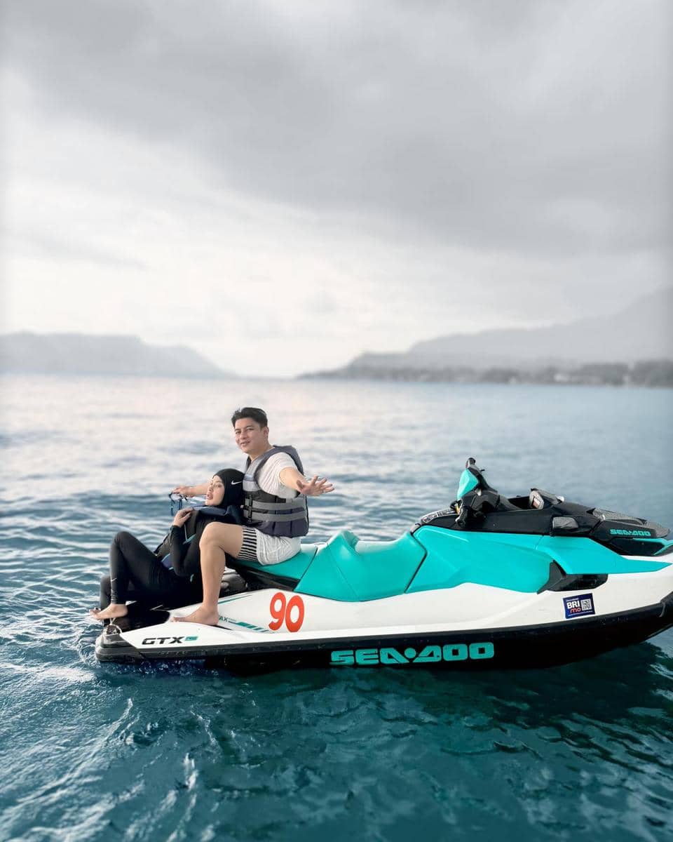 Sepasang suami istri menikmati waktu bersama di atas jet ski berwarna putih dan hijau toska di tengah laut dengan latar pegunungan.