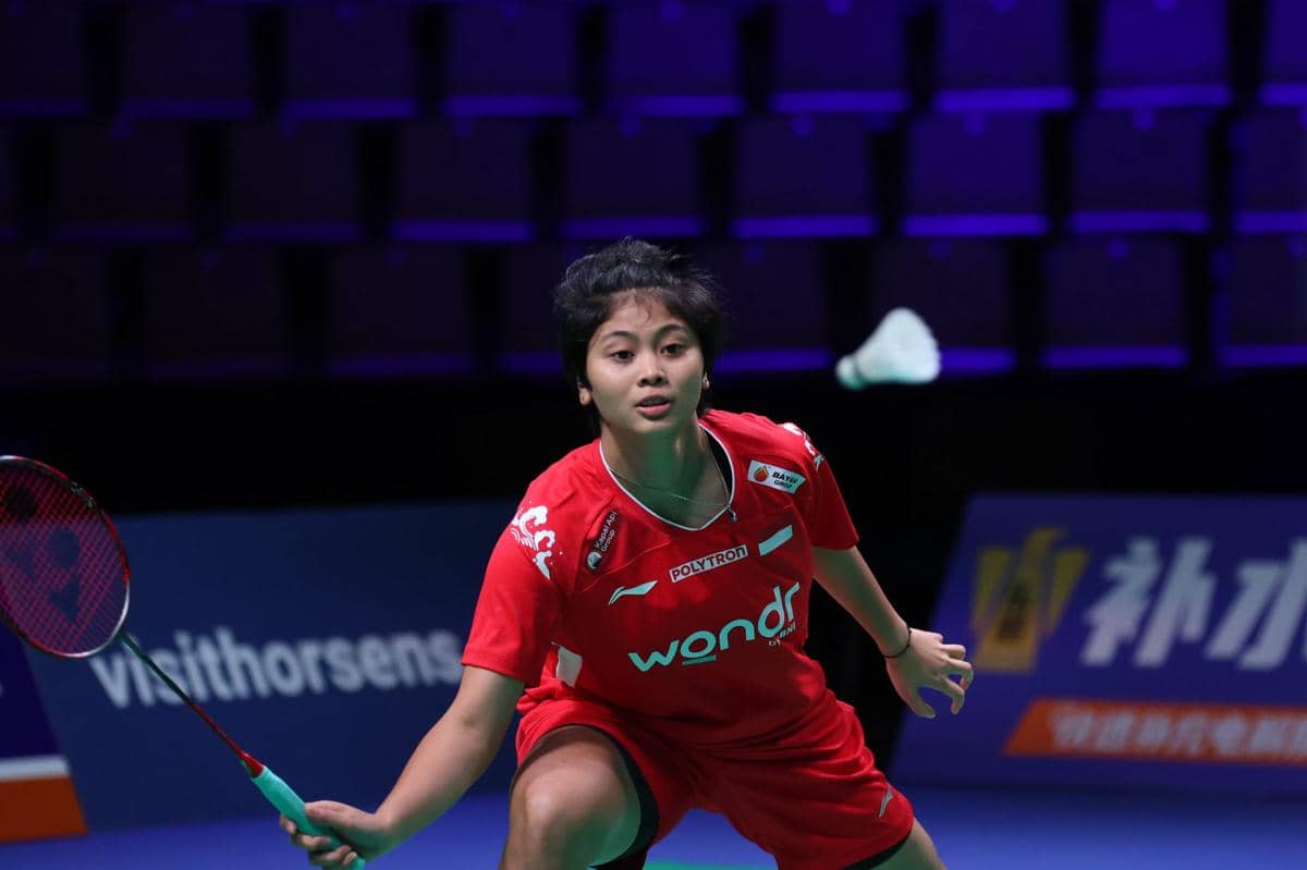 Seorang atlet bulu tangkis mengenakan seragam merah sedang fokus memukul shuttlecock di lapangan indoor dengan pencahayaan ungu.