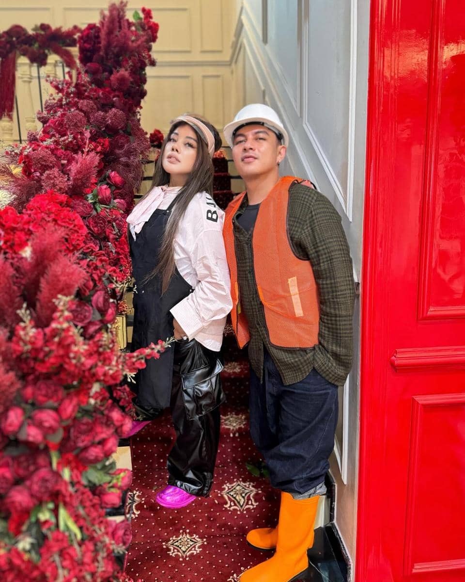 Sepasang pria dan wanita berpose di tangga berhias bunga merah, mengenakan pakaian kasual dengan rompi oranye dan sepatu bot warna cerah.