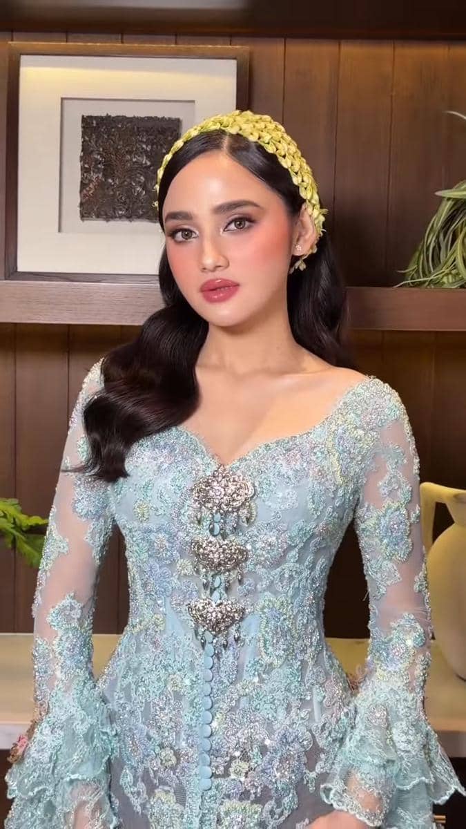Seorang perempuan mengenakan kebaya biru muda berhias payet dan bros perak, dengan rambut bergelombang serta hiasan kepala kuning.