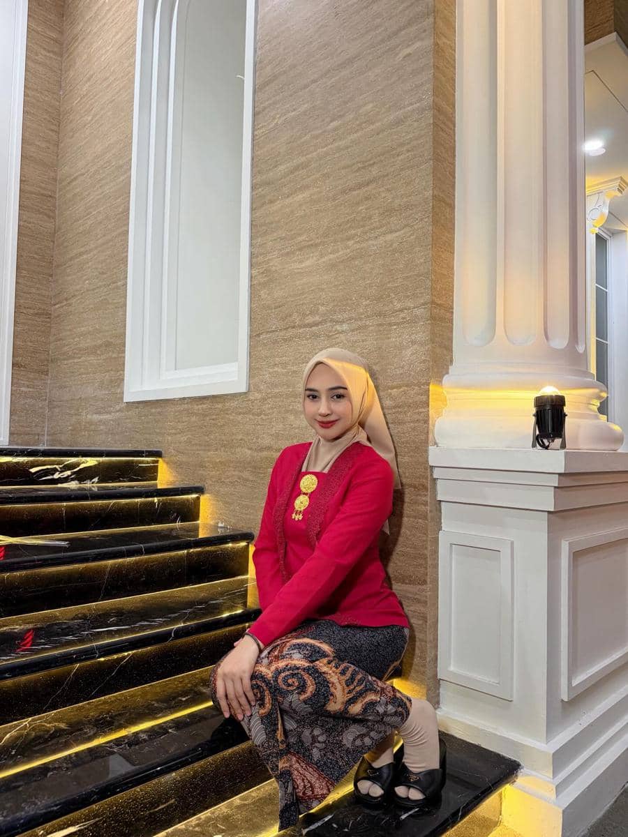 Seorang wanita berhijab mengenakan kebaya merah dan kain batik duduk di tangga marmer hitam dengan pencahayaan kuning hangat.
