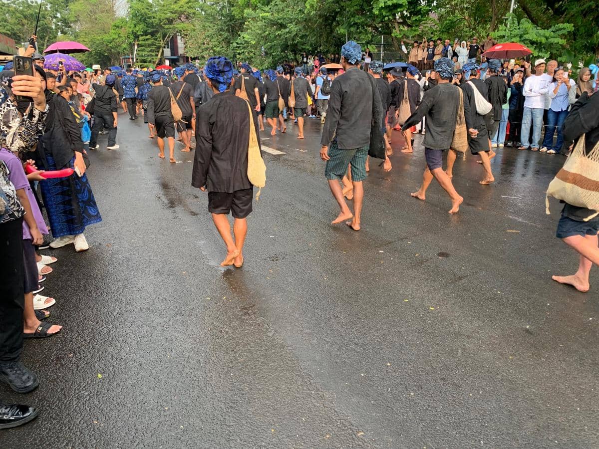 Warga adat Baduy berjalan kaki tanpa alas kaki mengenakan pakaian hitam dan ikat kepala biru dalam tradisi Seba di jalan yang basah.