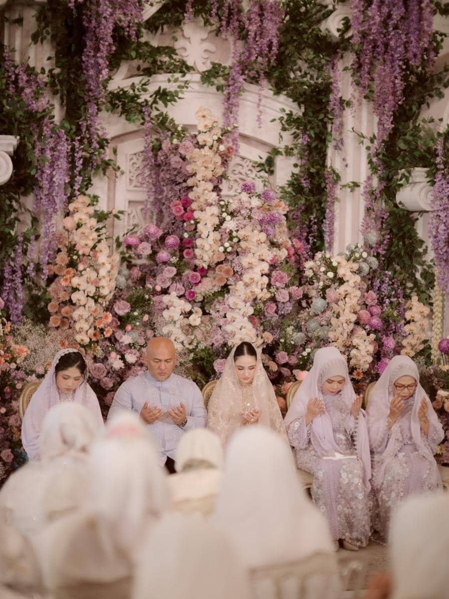 Keluarga mengenakan busana bernuansa ungu dan krem sedang berdoa bersama dalam acara pengajian dan siraman dengan dekorasi bunga yang indah.