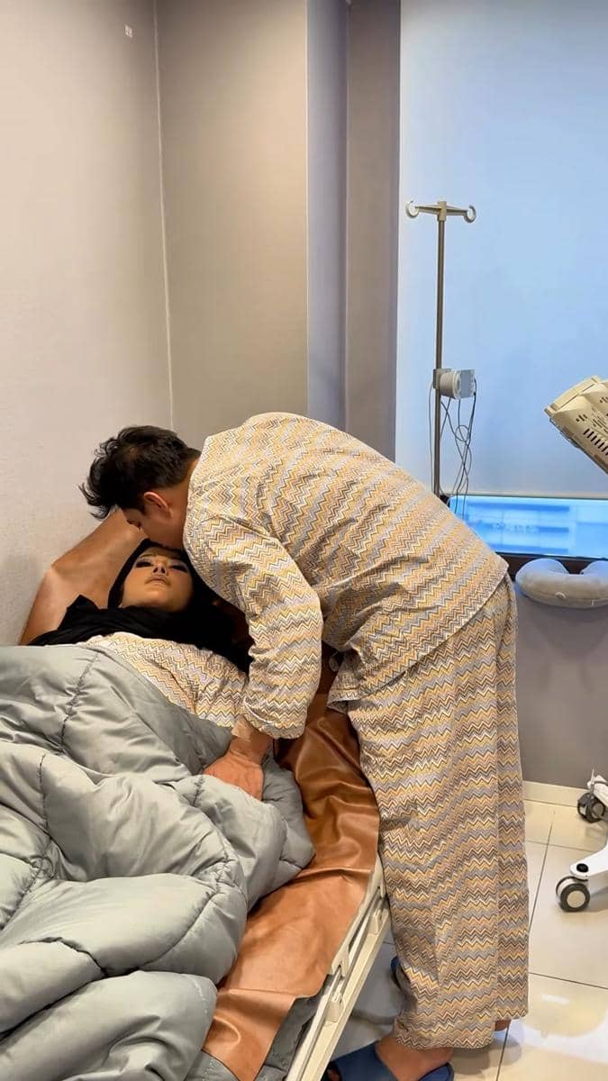 Seorang pria mencium kening wanita yang terbaring di tempat tidur rumah sakit, keduanya mengenakan piyama bermotif serupa.