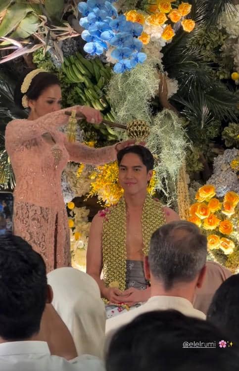 Seorang wanita mengenakan kebaya warna peach sedang melakukan prosesi siraman kepada seorang pria yang duduk dengan selendang bunga melati di bahunya.