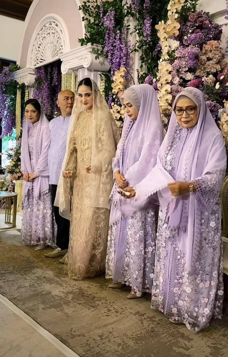 Keluarga mengenakan busana seragam ungu dan emas berpose di acara pengajian dan siraman dengan dekorasi bunga ungu dan putih.