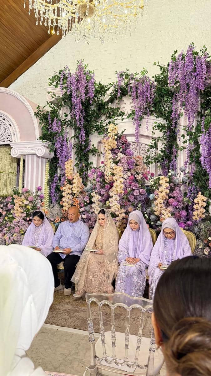 Suasana pengajian dan siraman menjelang pernikahan dengan dekorasi bunga ungu dan pastel, dihadiri keluarga yang mengenakan busana senada.