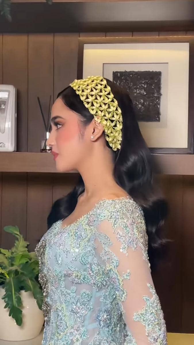 Seorang perempuan mengenakan kebaya biru muda berhias payet dan sanggul dengan hiasan melati di kepala, berdiri di dalam ruangan kayu.