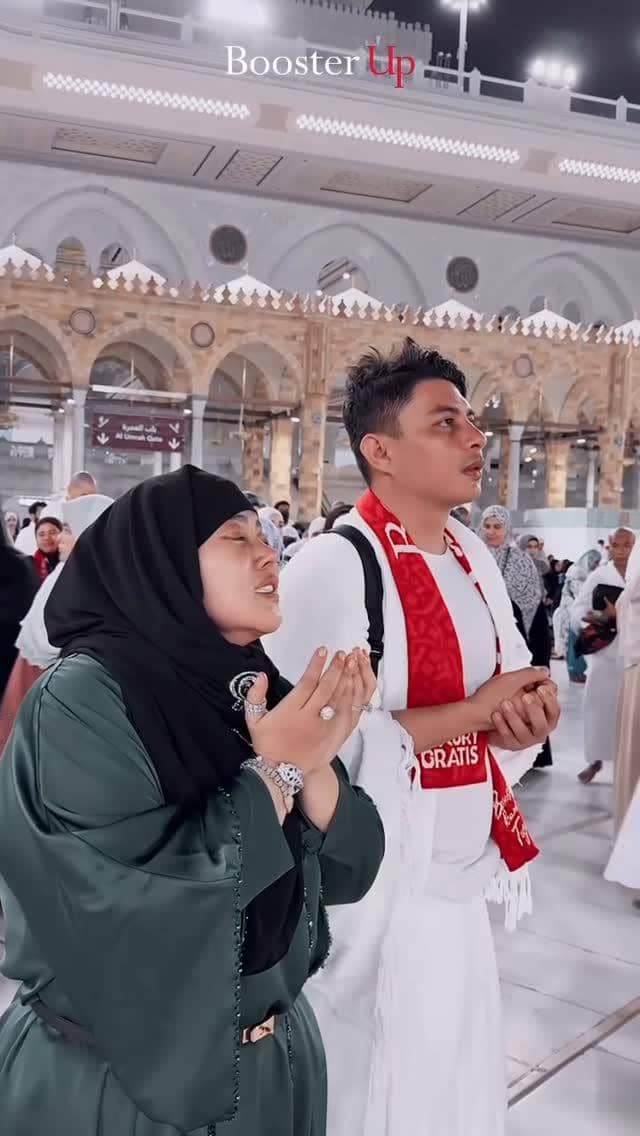 Sepasang suami istri mengenakan pakaian ihram dan abaya hijau sedang berdoa khusyuk di area Masjidil Haram pada malam hari.