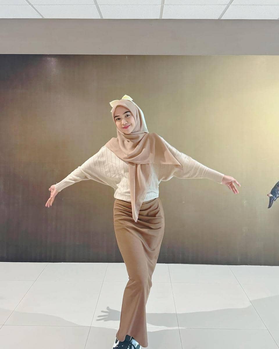 Seorang perempuan berhijab berpose dengan tangan terbuka di ruangan bercat cokelat, mengenakan sweater krem dan rok panjang cokelat.