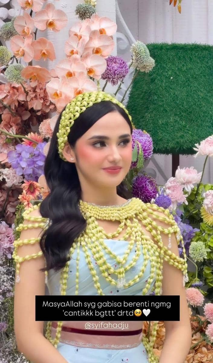 Seorang perempuan mengenakan kebaya biru muda berhias rangkaian melati di acara siraman, dengan latar bunga warna-warni yang indah.