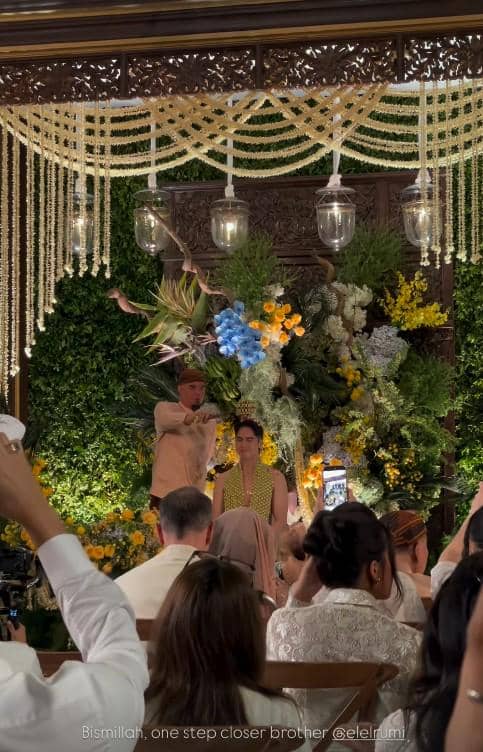 Seorang pria mengenakan pakaian tradisional melakukan prosesi siraman kepada seorang wanita di acara pengajian dengan dekorasi bunga dan janur.