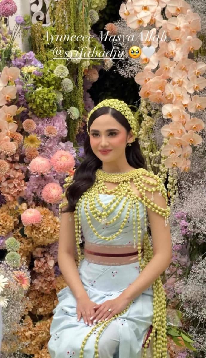 Seorang perempuan mengenakan kebaya biru muda dengan hiasan melati di kepala dan tubuh, duduk di depan dekorasi bunga warna-warni saat acara siraman.