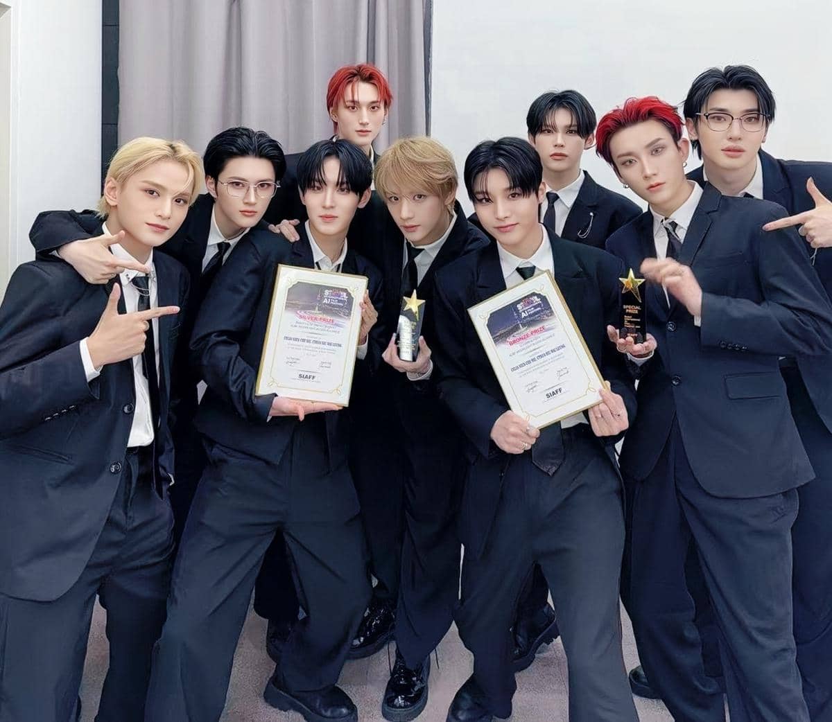 Sembilan anggota grup musik mengenakan setelan jas hitam memegang piala dan sertifikat penghargaan di sebuah ruangan.