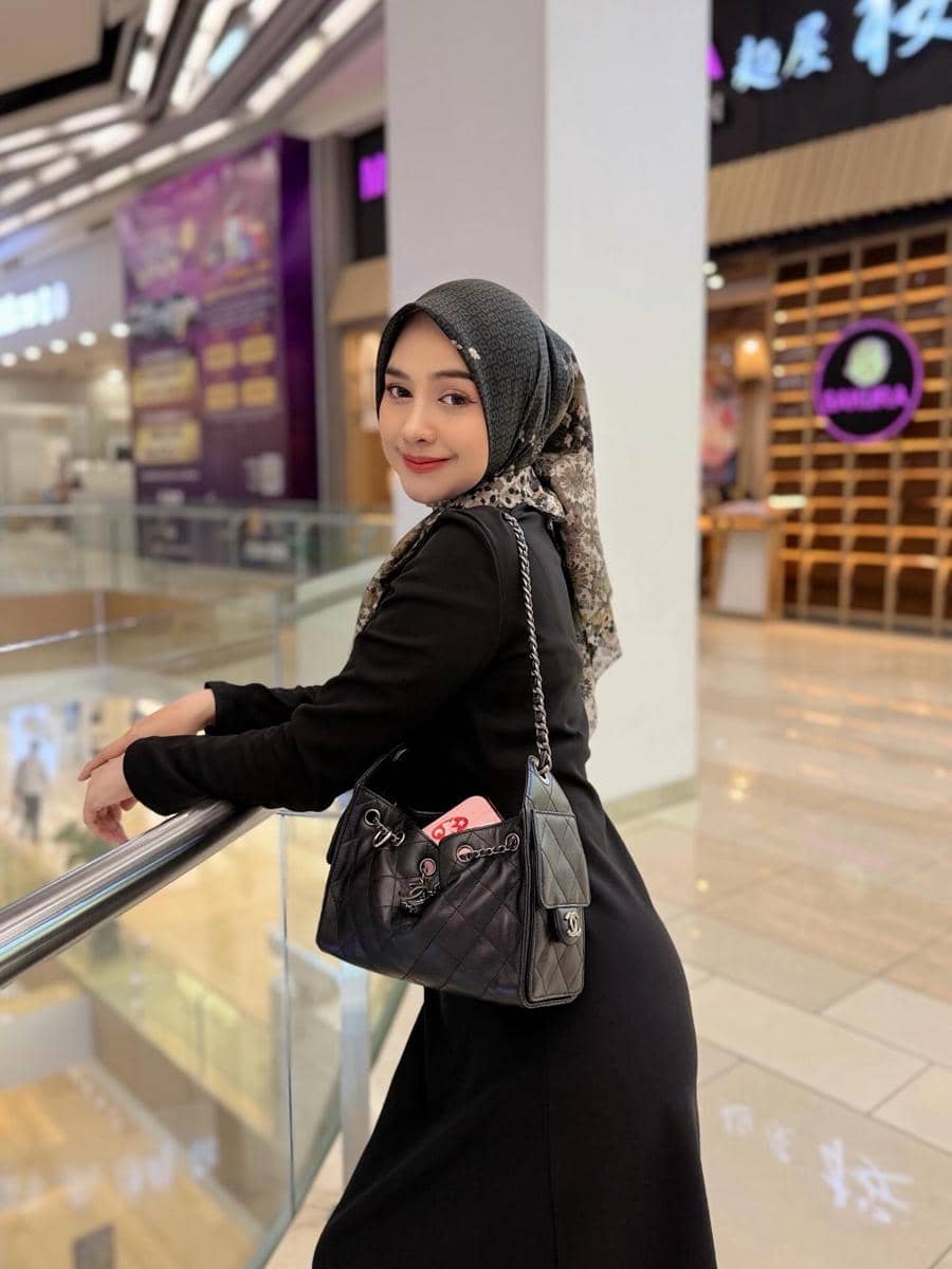 Seorang wanita berhijab mengenakan busana hitam dan tas kulit hitam berdiri di area pusat perbelanjaan modern.