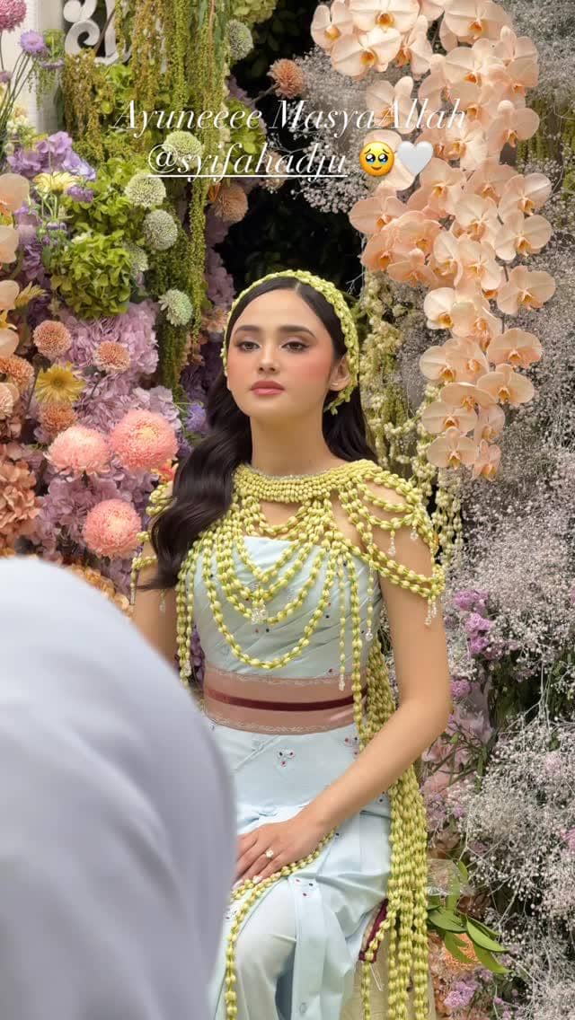 Seorang perempuan mengenakan kebaya biru muda dan hiasan melati duduk di depan dekorasi bunga warna pastel pada acara pengajian dan siraman.