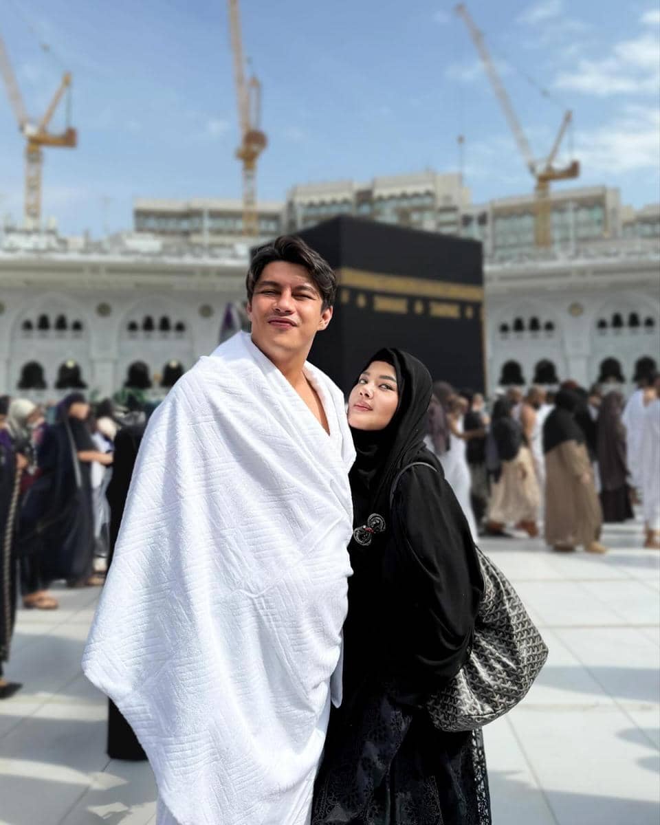 Sepasang suami istri berpose di depan Ka'bah di Masjidil Haram, mengenakan pakaian ihram dan abaya hitam dengan latar suasana ibadah haji.