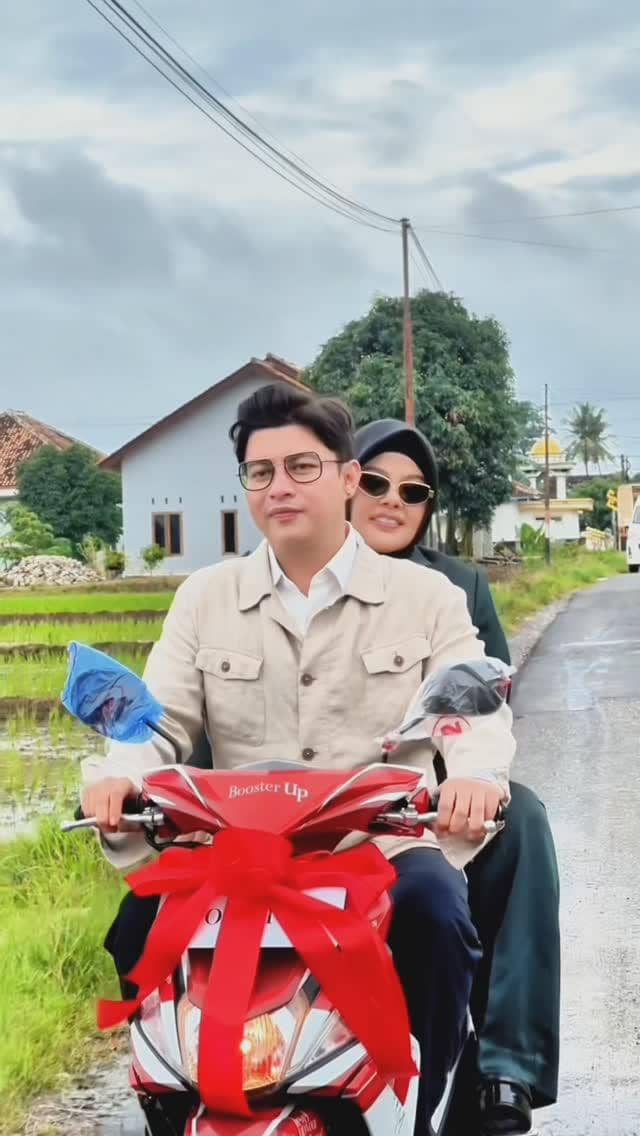Sepasang suami istri berkendara motor berhiaskan pita merah di jalan pedesaan dengan latar rumah dan sawah hijau.