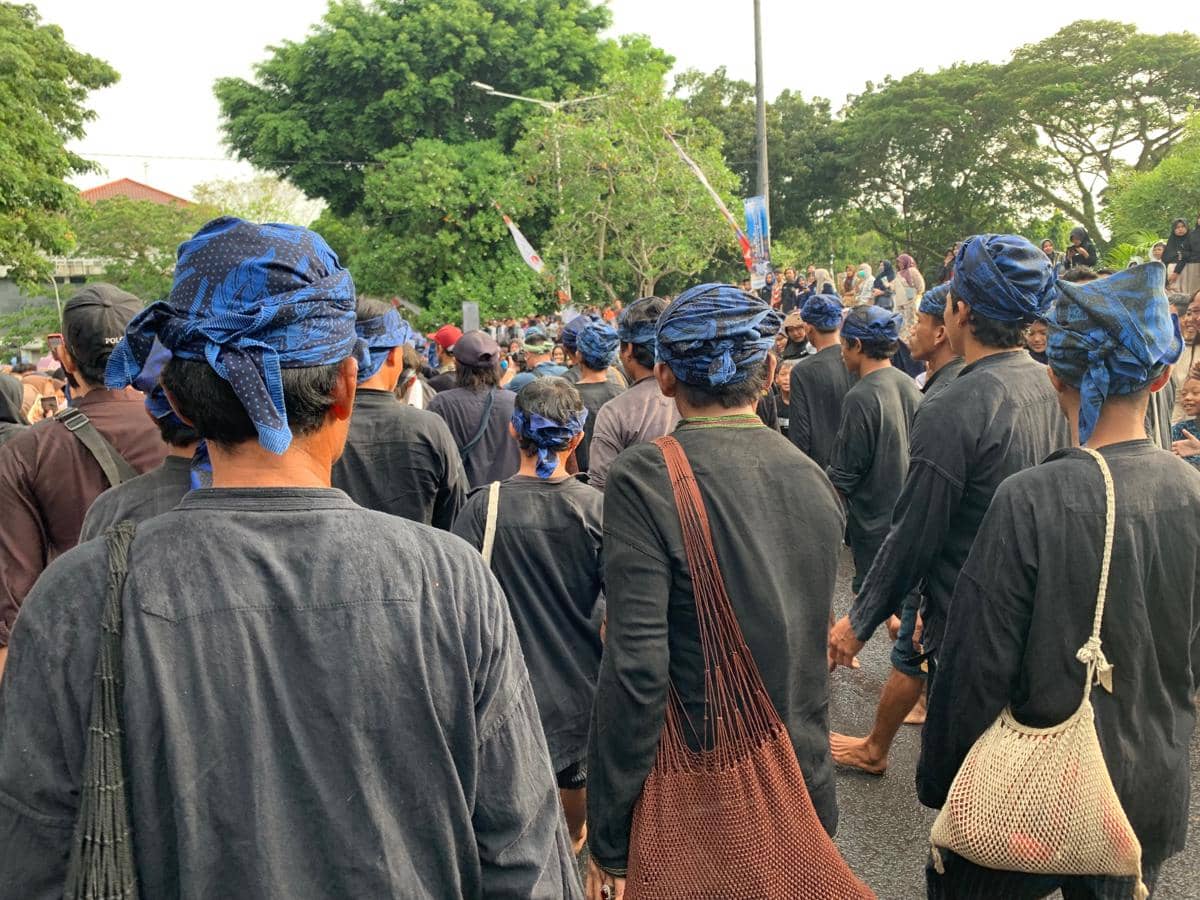 Warga adat Baduy mengenakan pakaian hitam dan ikat kepala biru berjalan bersama dalam tradisi Seba di ruang terbuka.